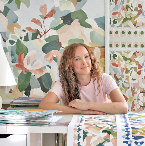 A woman with long curly light brown hair sits at a white table in front of a large colorful floral artwork and patterned prints. She is wearing a light pink t-shirt and looking to the side.