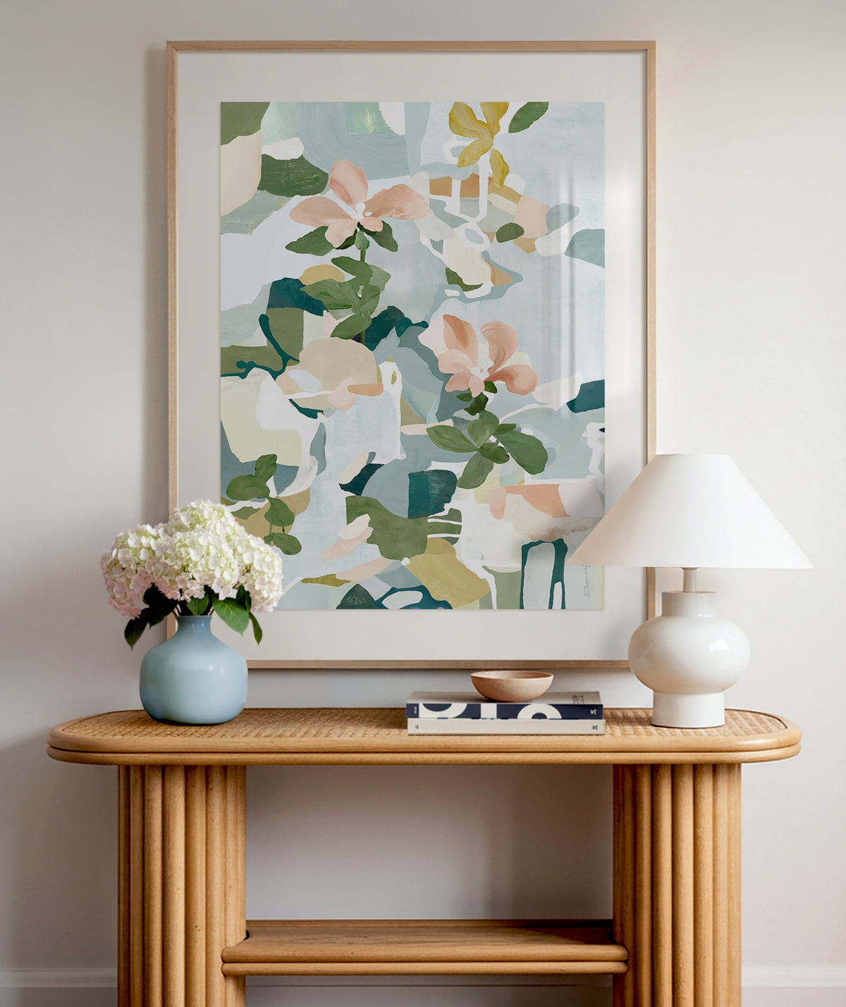 An abstract floral art print in light blue, pink, green and yellow displayed framed above a wooden entryway table with a white lamp and blue vase with white flowers.