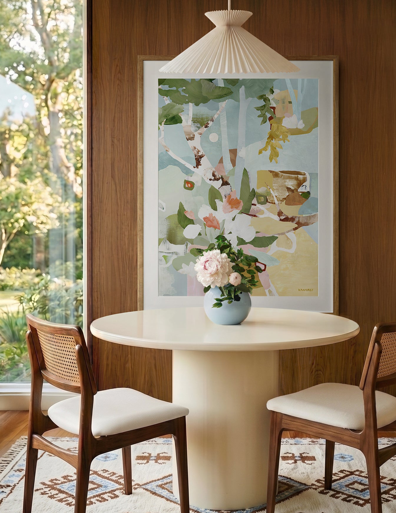 The art print titled Evening Hideaway depicting a semi-abstract wild garden hangs framed and matted on a wood-paneled wall above a round cream pedestal dining table with two wooden chairs. A blue vase with pink flowers sits on the table, and a window on the left shows green outdoor foliage.