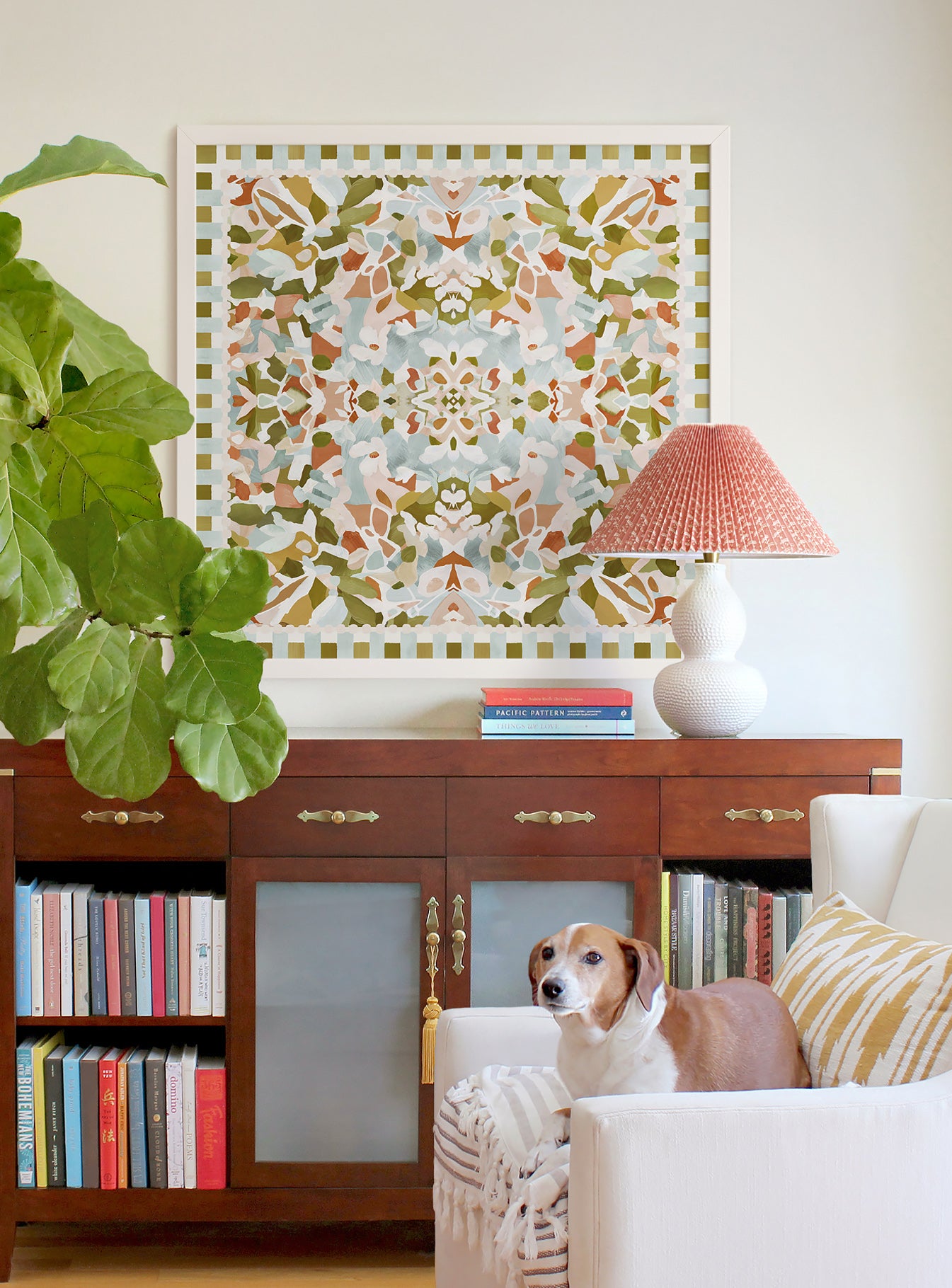 Large square, abstract symmetrical art in olive green, light pink, light soft blue, and terracotta colors displayed above a bookcase cabinet. There is a dog lying on a white accent chair.
