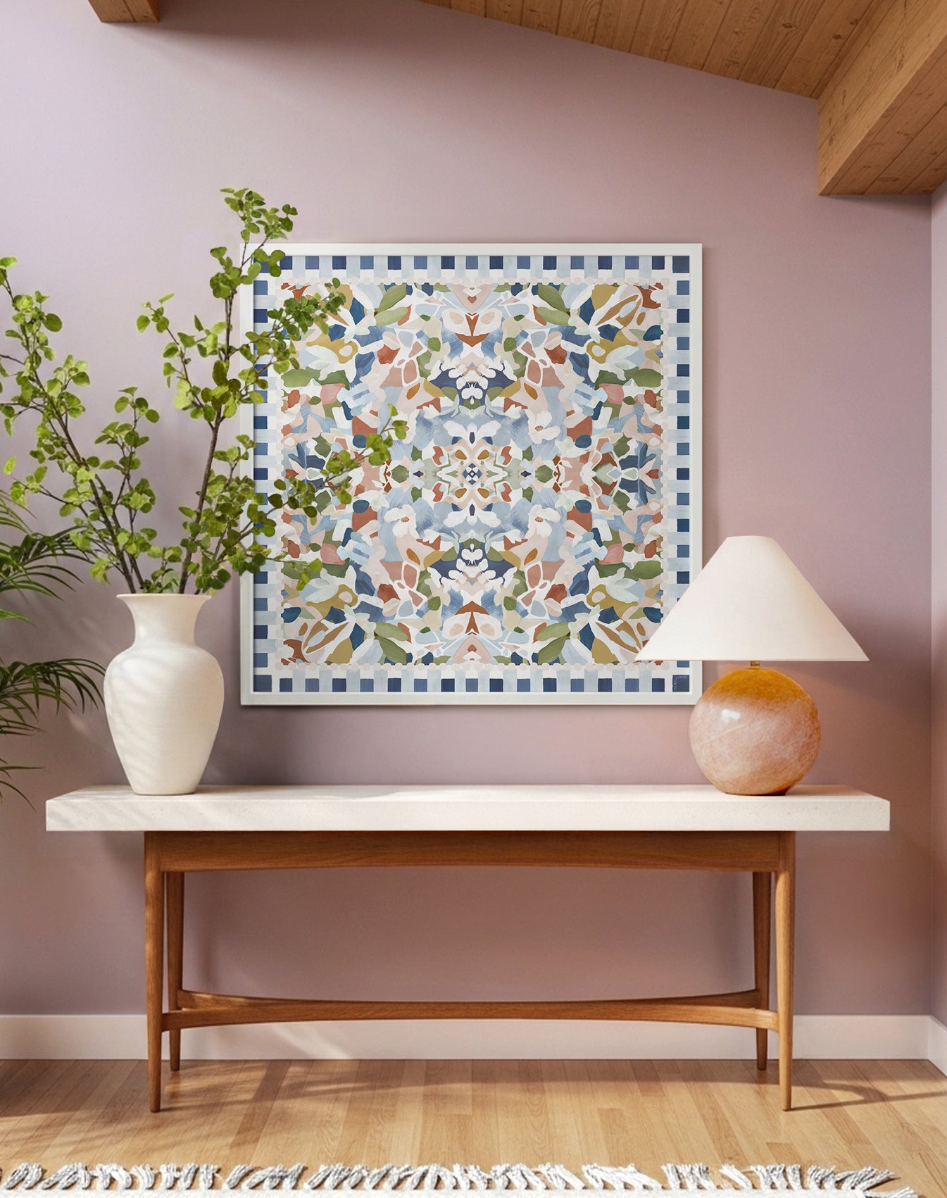 Large square, abstract and floral art print featuring blue, green, light pink, and terracotta colors displayed in a white frame above an entryway table with a vase and a pink lamp.