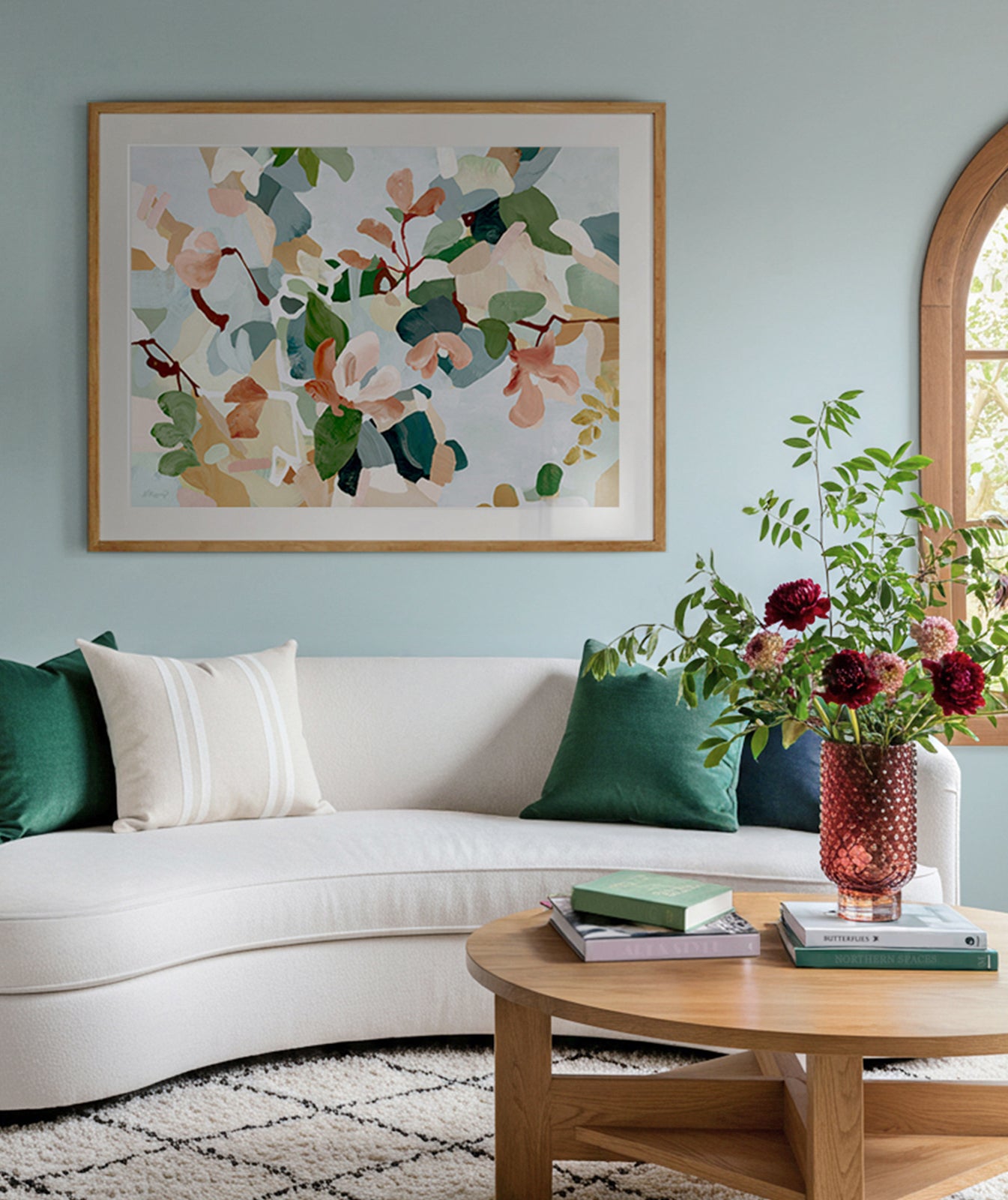Living room with a white sofa, decorative pillows, a coffee table with a vase and books, and a large framed abstract floral painting in blues, soft reds and greens.