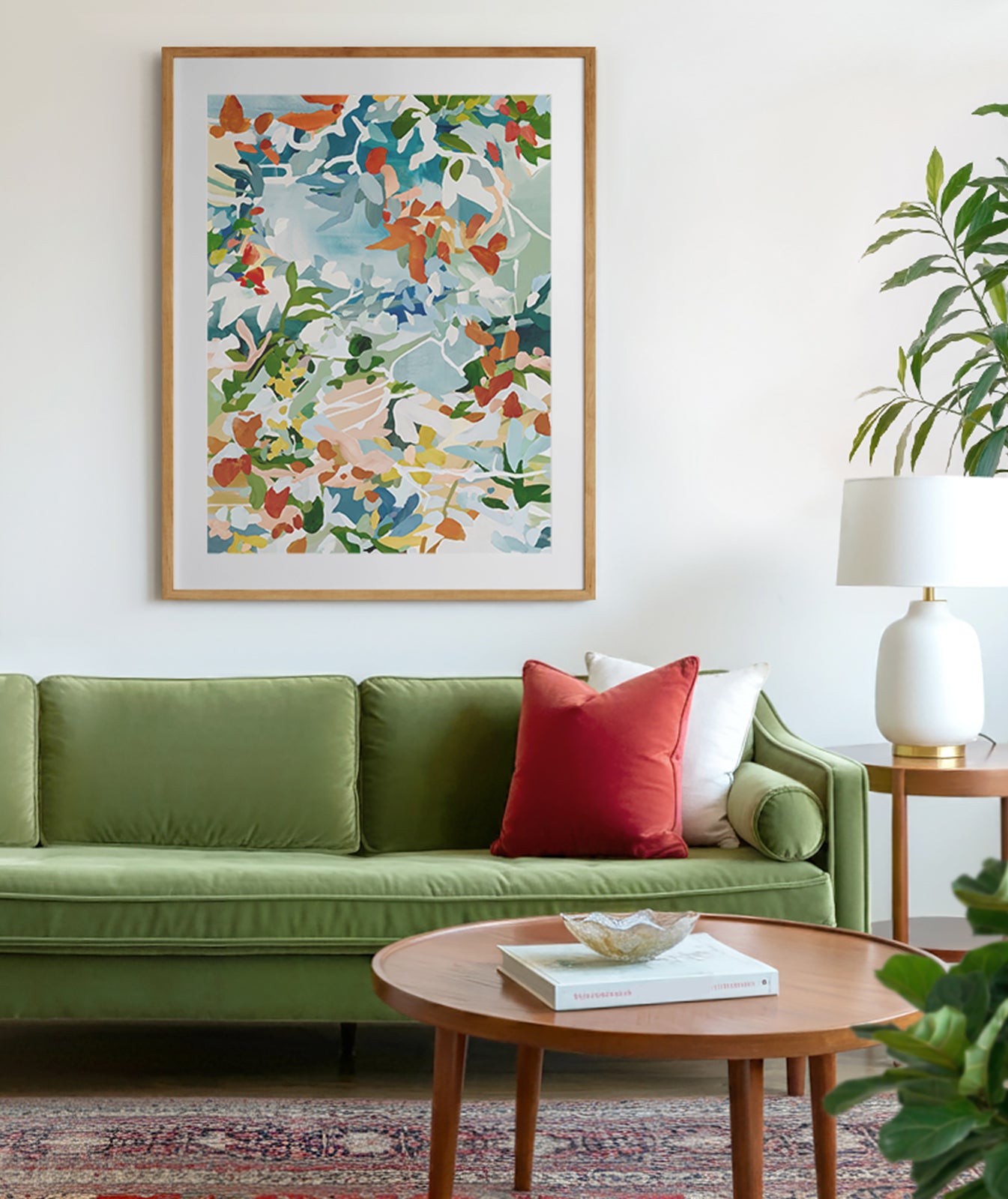 A mid century modern living room with velvet green sofa and red and white cushions, there is a traditional red carpet and round, wooden coffee table. The art is a floral abstract with warm reds, greens and blues.