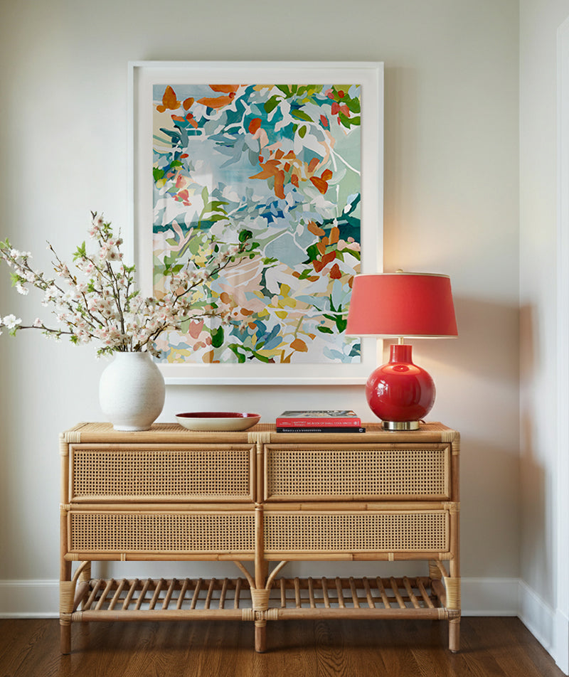 Bright entryway decor featuring a rattan console table, red modern lamp, cherry blossom white vase, abstract art in bright floral theme, the art is framed in white. The overall feel is feminine modern cottage charm, a warm and inviting interior design style that feels traditional yet fresh.