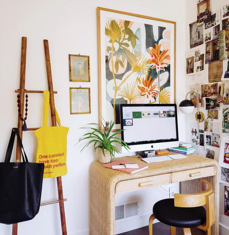 A colorful desk space with a computer and colorful art above it, various photos on the wall next to it, as well we a decorative ladder with bags hanging on it.