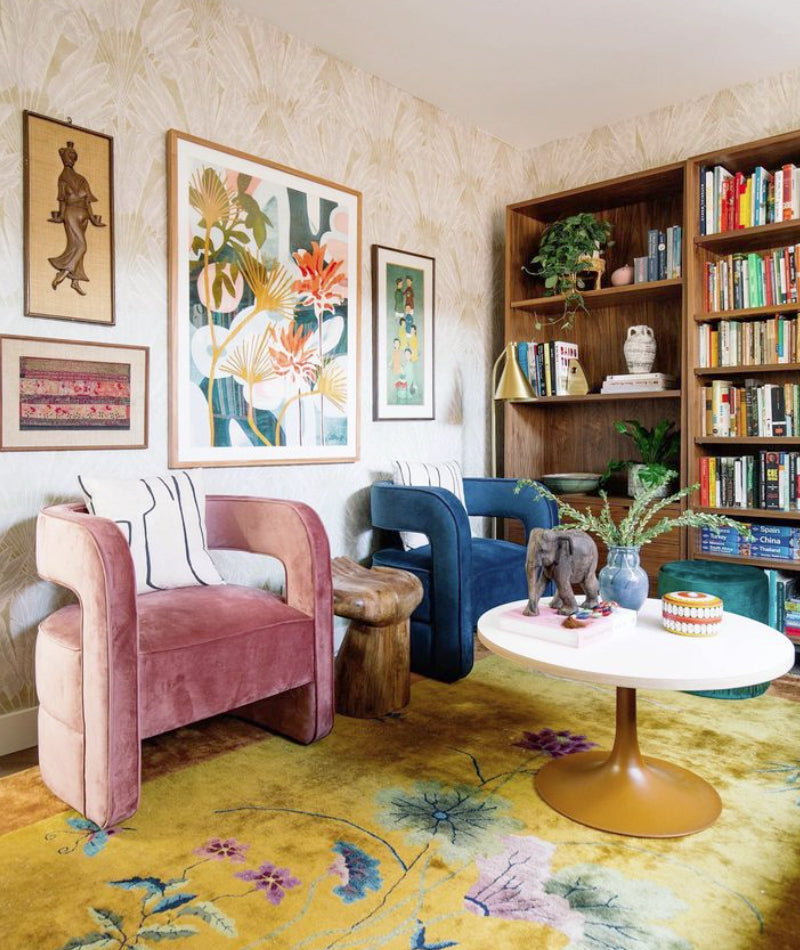 A colorful office space featuring a pink accent chair and a blue accent chair, a yellow rug and several pieces of art on the wall. There is a bookcase with colorful books along one wall and a round, white coffee table.