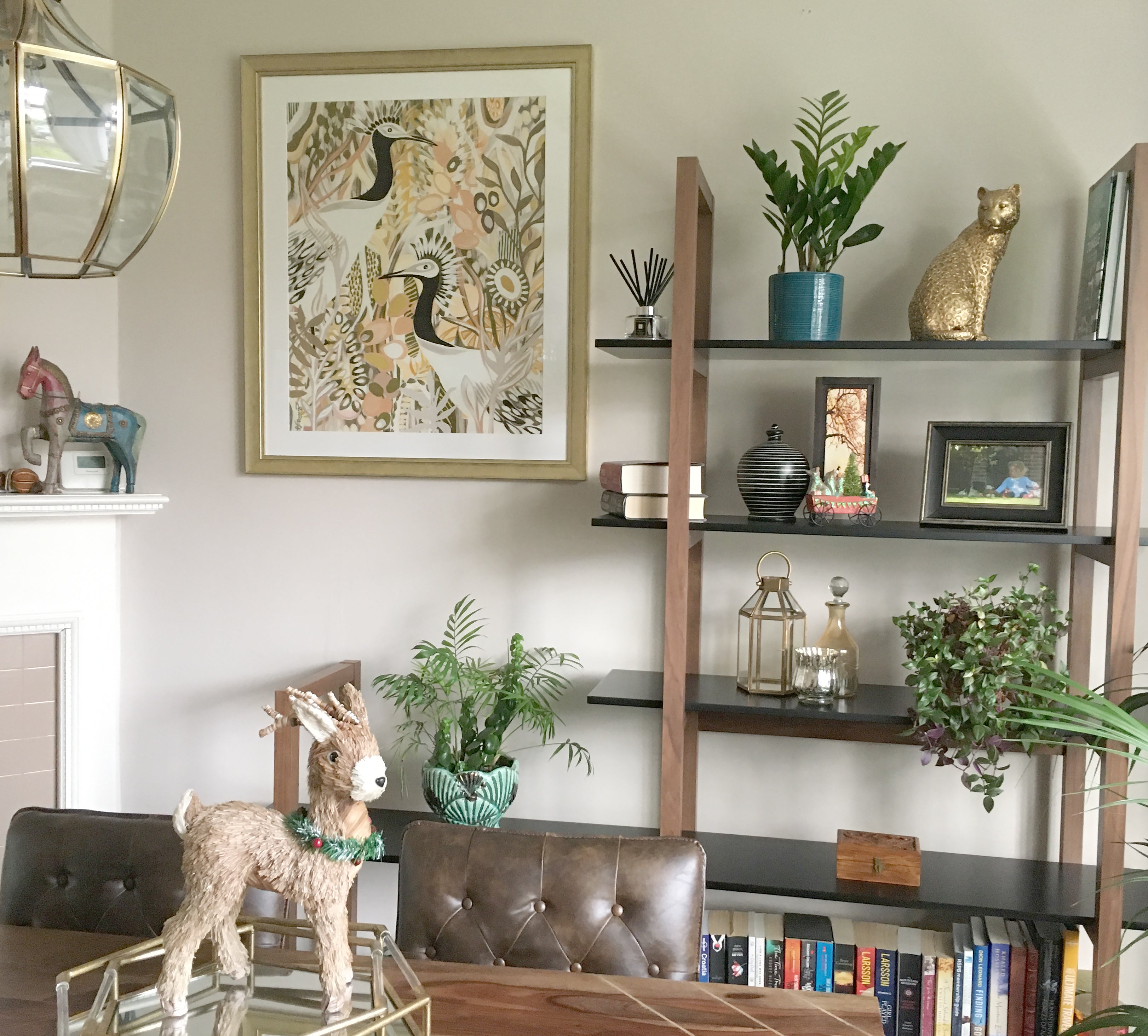 A close-up of a neutral dining room, there is a shelf along one wall with various decor items on it. There is one framed art piece on the wall featuring two birds in a colorful garden. A glass pendant light hangs above a wooden table.