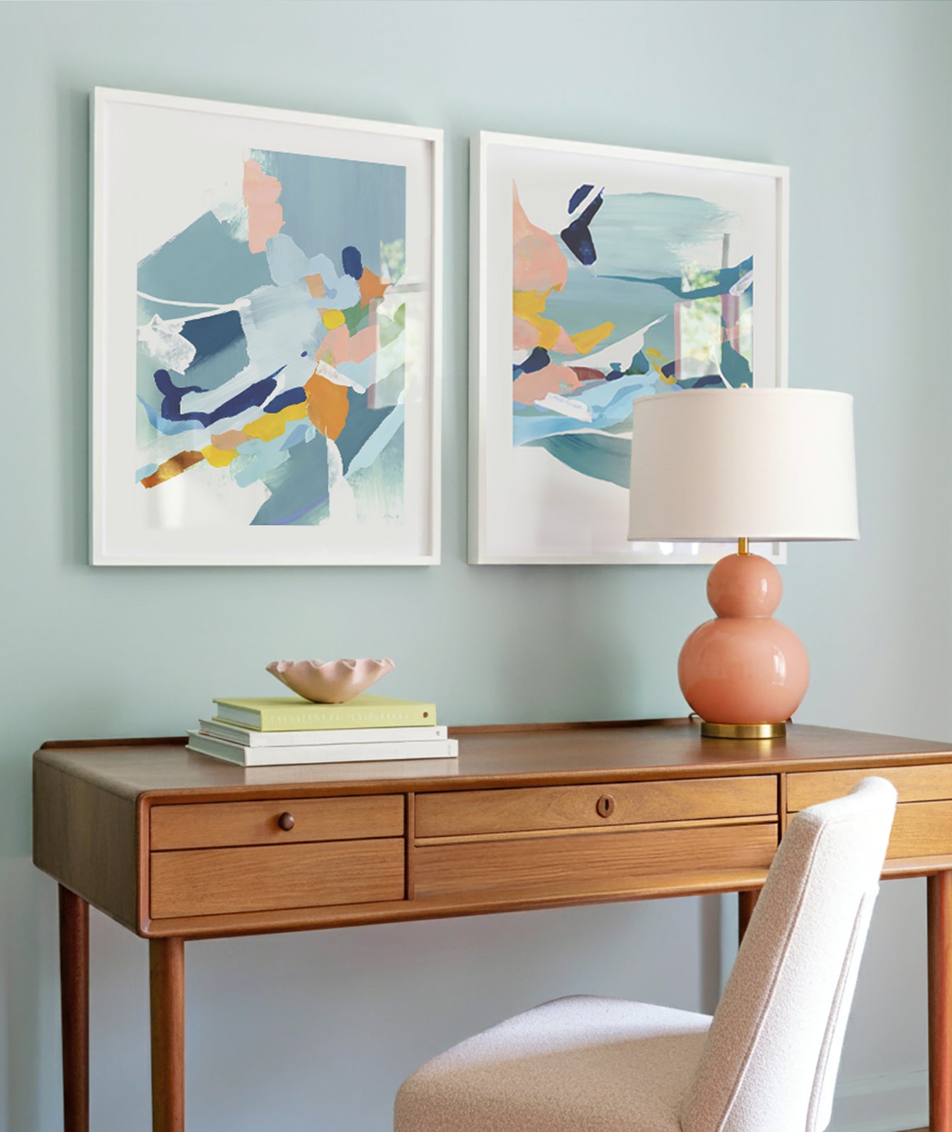 A desk space with a mid century modern, wooden desk, there is a modern round lamp in a light coral color on the desk, the wall color is light blue. A modern chair in a light pink fabric is in front of the desk, there are books and two matching, abstract art print with blues, pinks, yellow and orange colors framed in white above the desk.