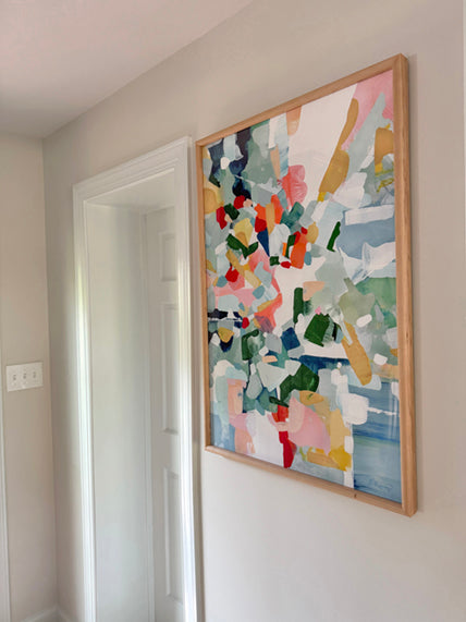 Customer photo of  the artwork titled "Positano" consisting of a colorful mix of blues, pinks, yellows and greens in an abstract style. The art is framed in a light wood frame displayed in a hallway.