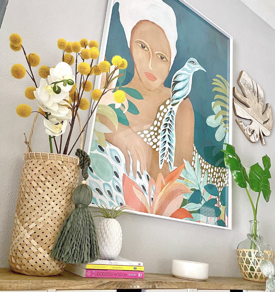 Customer photo of  the artwork titled "Lounging" which features a woman in a colorful garden. The art is framed in a white modern frame and displayed in a bright entryway above a white cabinet.