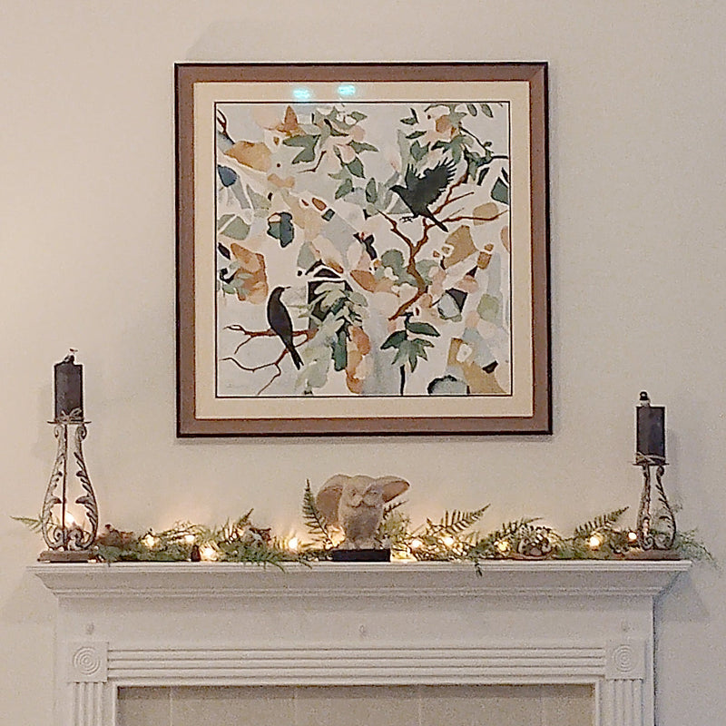 A framed wall art print featuring a bird and leaf pattern hangs above a white fireplace mantel decorated with candles, greenery, and a small elephant figurine. The scene is set in a living room with a beige sofa visible at the bottom.