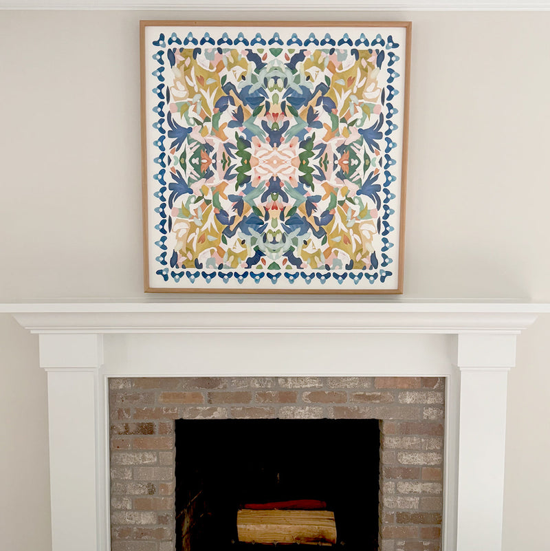 The square artwork titled Reverie  featuring blue, yellow, pink and green abstract patterns hangs centered above a white fireplace mantel and brick hearth.