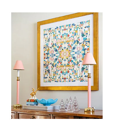 A framed colorful abstract art print with a gold frame hangs on a beige wall above a wooden console table. The print features a colorful geometric pattern with blue, yellow, red, and green shapes on a white background.