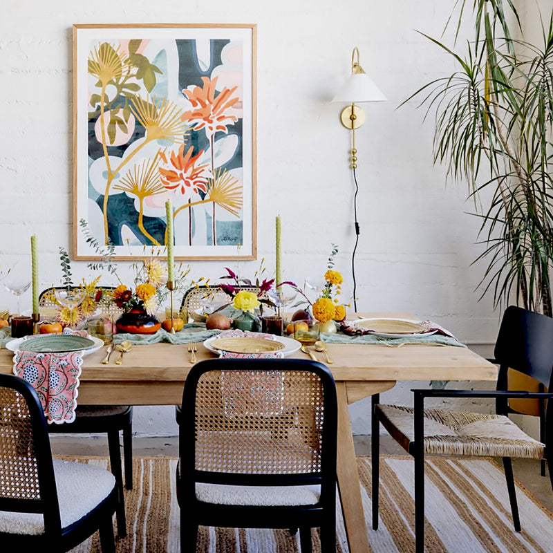 A styled dining room scene by interior designer Dabito showcasing a light wood dining table set for a meal, surrounded by black chairs. The Mykonos art print featuring botanical coral, red and dark blue design hangs on the white wall above the table.