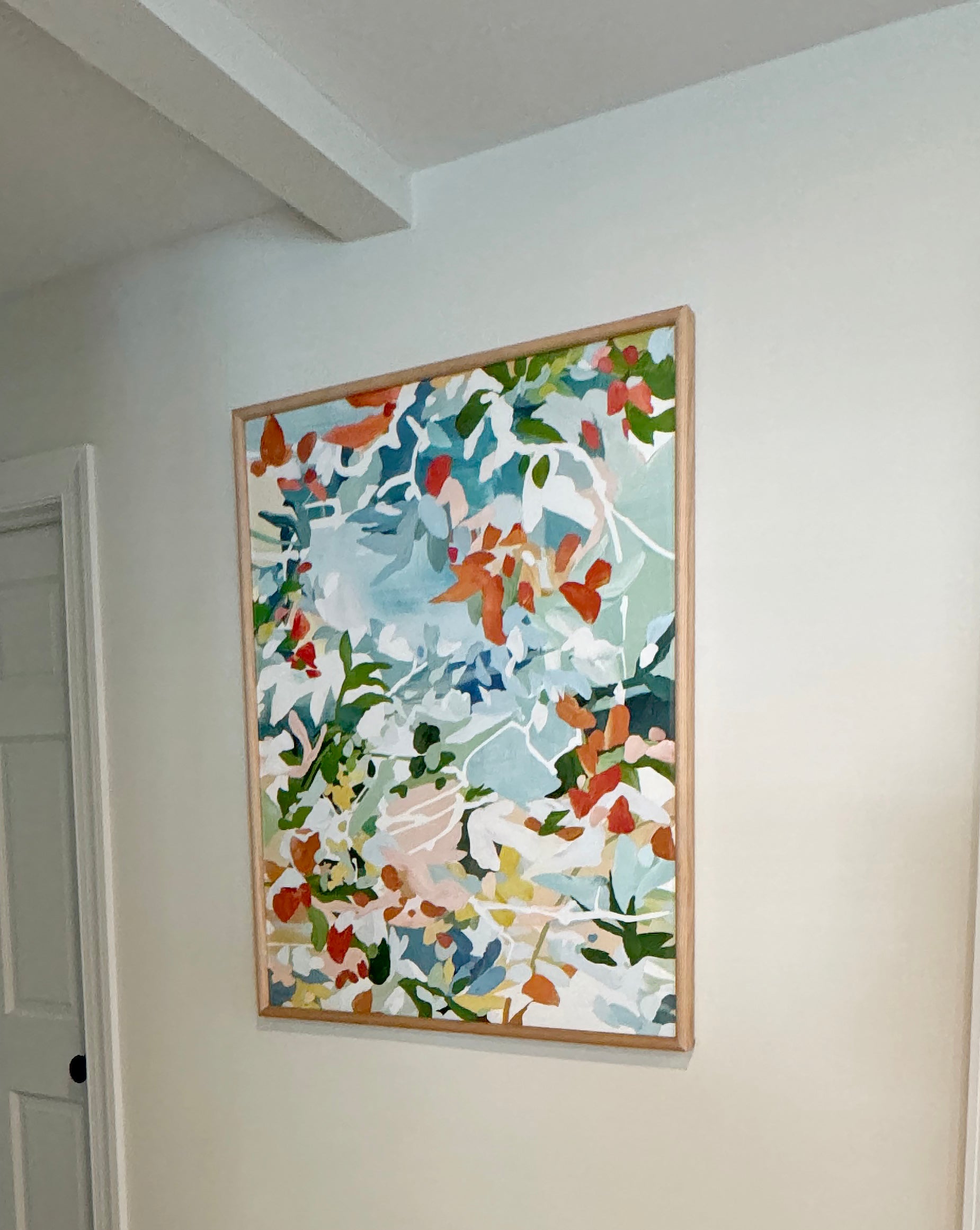 Customer photo of the abstract floral art titled "Agosto" featuring a blue, red, green and pink color palette. The art is framed in a light wood frame and displayed in a hallway.