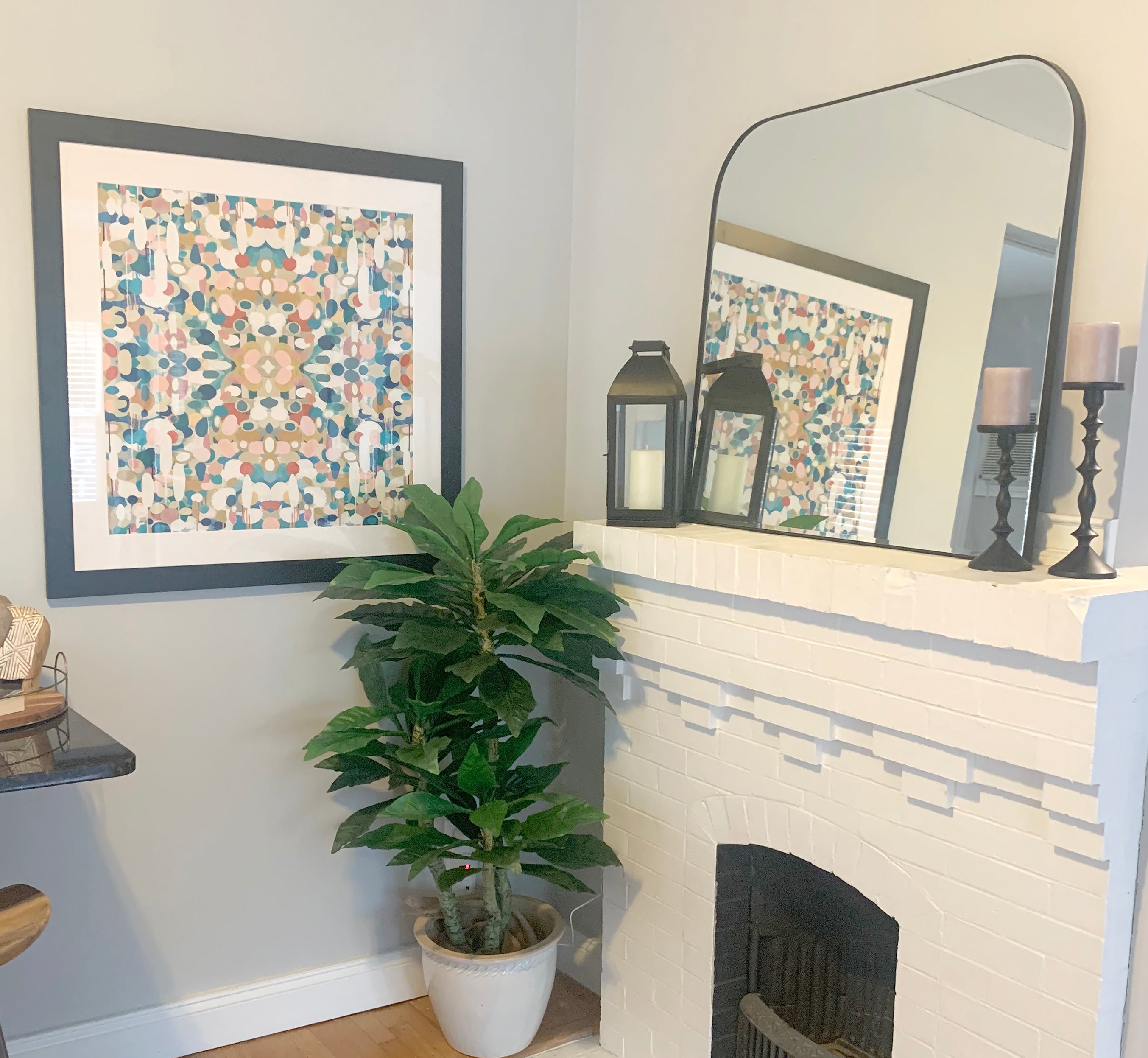 A close-up of a white fireplace with the artwork titled 'Euphoria' featuring various shades of blue, pinks and reds. The fireplace has a mirror, candle and and lantern on it, there is a plant of the floor.