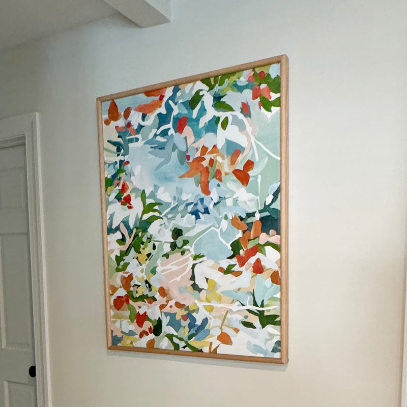 Artwork titled Agosto featuring a colorful abstract garden in red, blue, pink and green colors displayed in a wooden frame on a white wall.