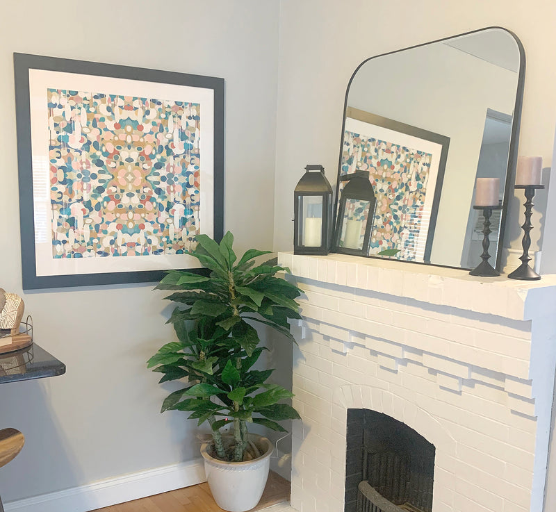 A styled interior corner displaying the artwork titled Euphoria featuring a colorful, symmetrical pattern with turquoise blues, reds and pinks framed on a light gray wall next to a white brick fireplace. A potted green plant sits below the frame, and a large black-framed mirror on the mantel reflects the room.
