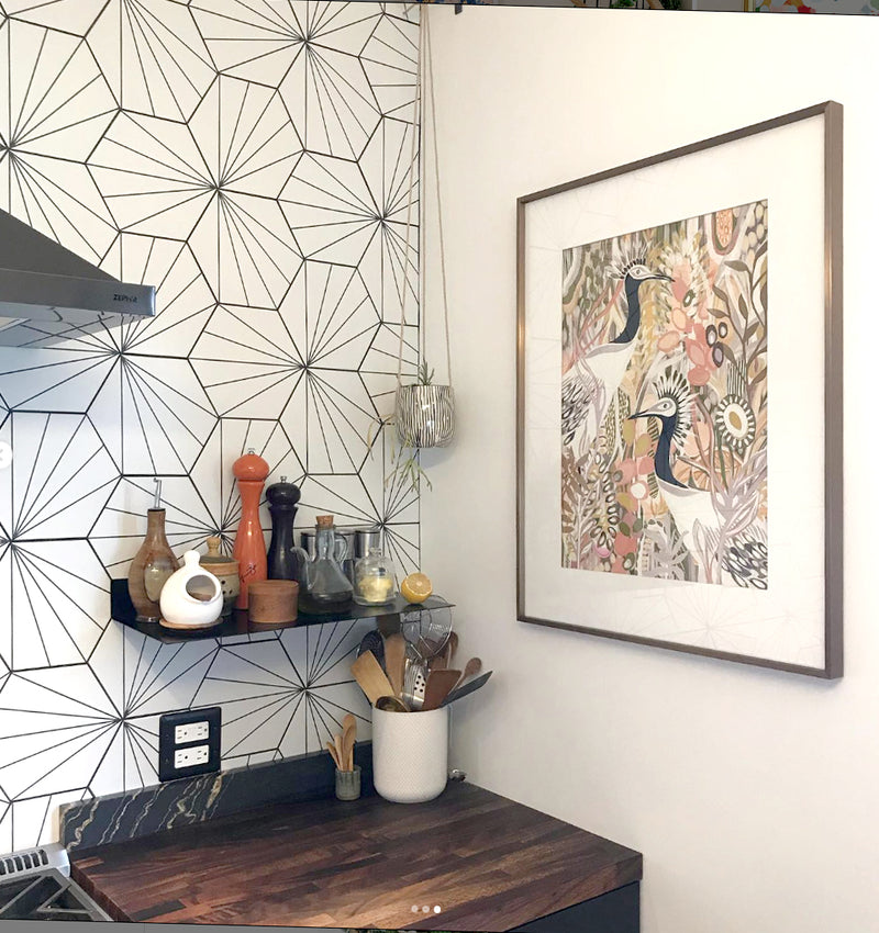 The artwork titled Garden Stroll featuring two white and black crane birds against a colorful garden featured in a kitchen corner with geometric wall tiles, a framed artwork, and various kitchen items.