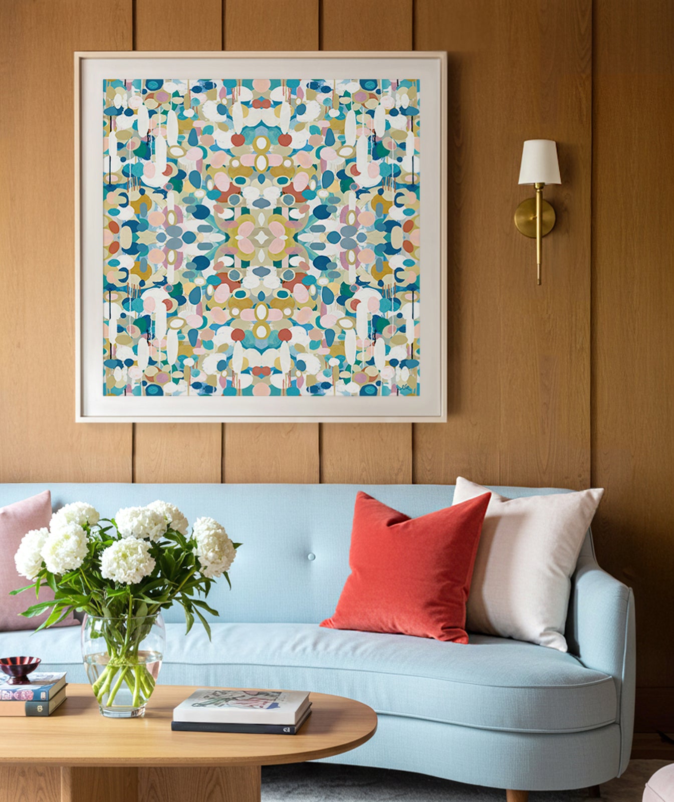 A colorful abstract fine art print with a kaleidoscopic design in turquoise blue, dark blue, red, pink and yellow tones framed and displayed above a light blue sofa against a wood paneled wall.