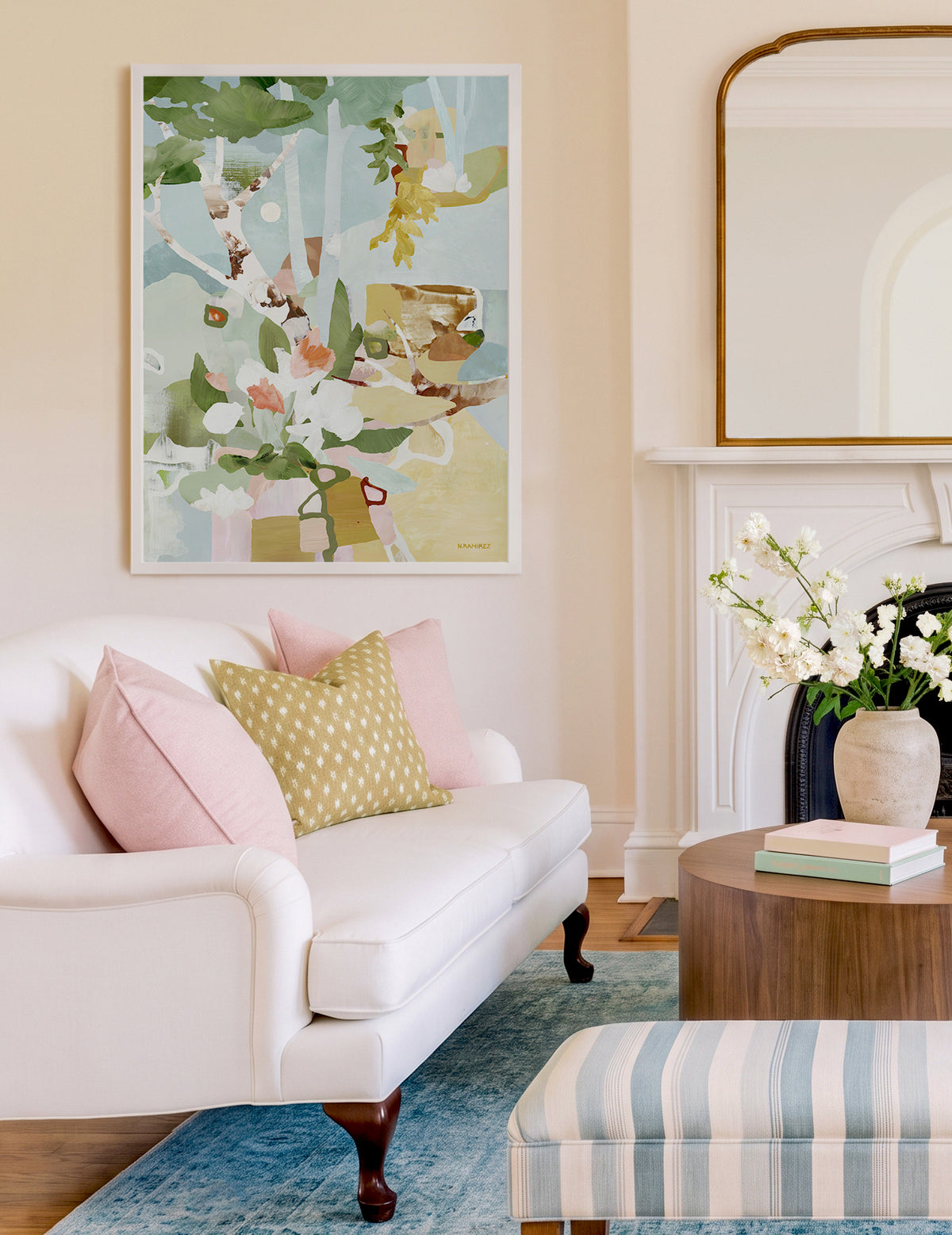 A framed fine art print titled Evening Hideaway depicting a semi-abstract, wild garden displayed hung on a warm white wall above a white sofa with pink and yellow pillows styled in a bright living room.