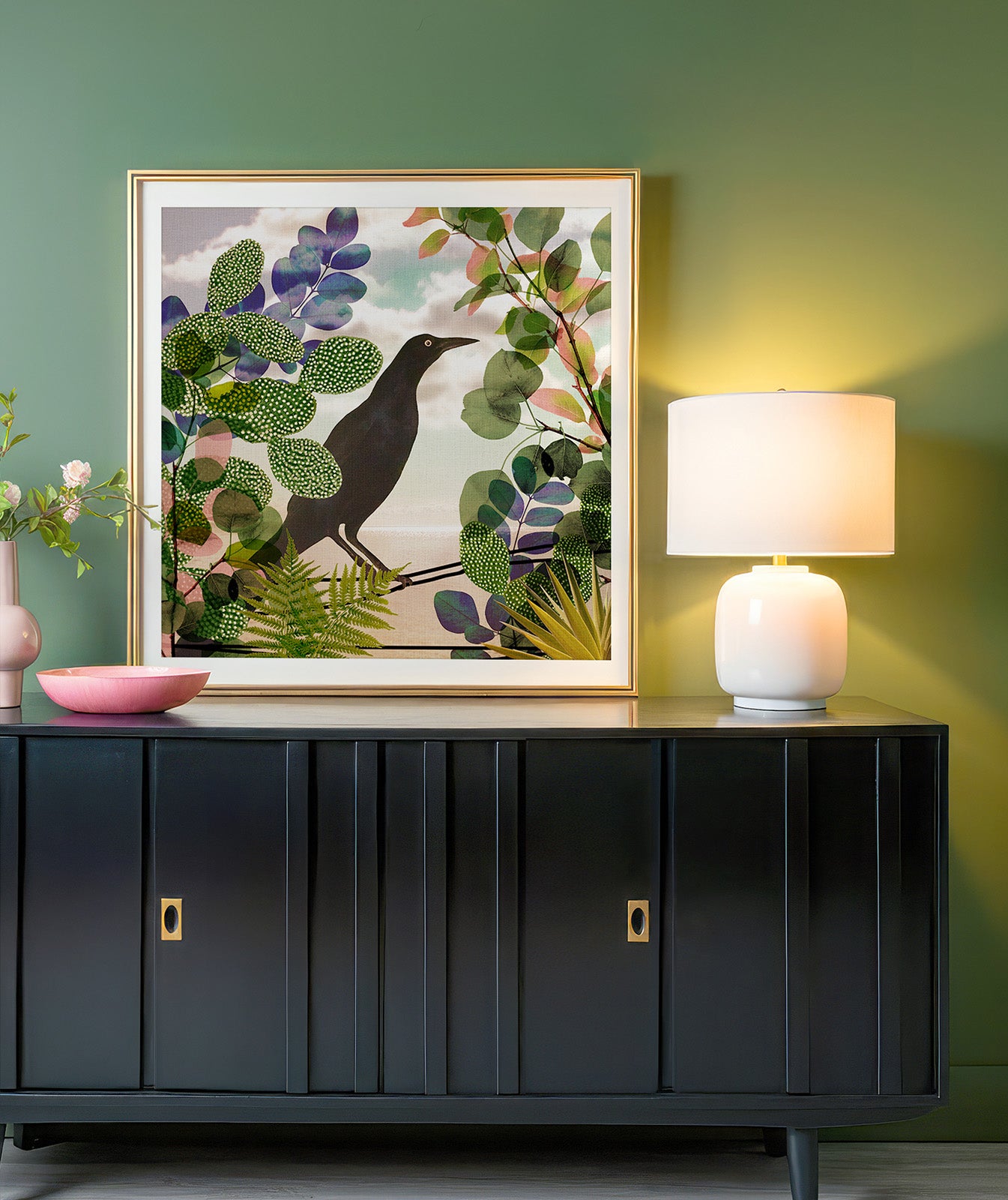 A black cabinet against a chartreuse wall with a modern white lamp on top. There is a large, square piece of art featuring a Nicaraguan Grackle bird surrounded by tropical plants.