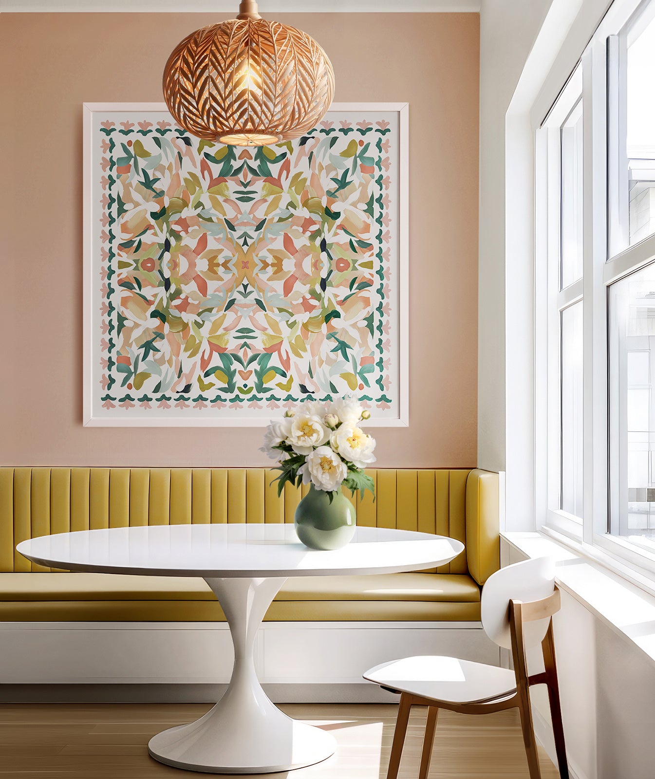 A modern kitchen with a muted pink wall, white, mid century modern round table, yellow banquette, there is a green vase with flowers on the table. A rattan pendant light hangs in front of a kaleidoscopic art print framed in white.