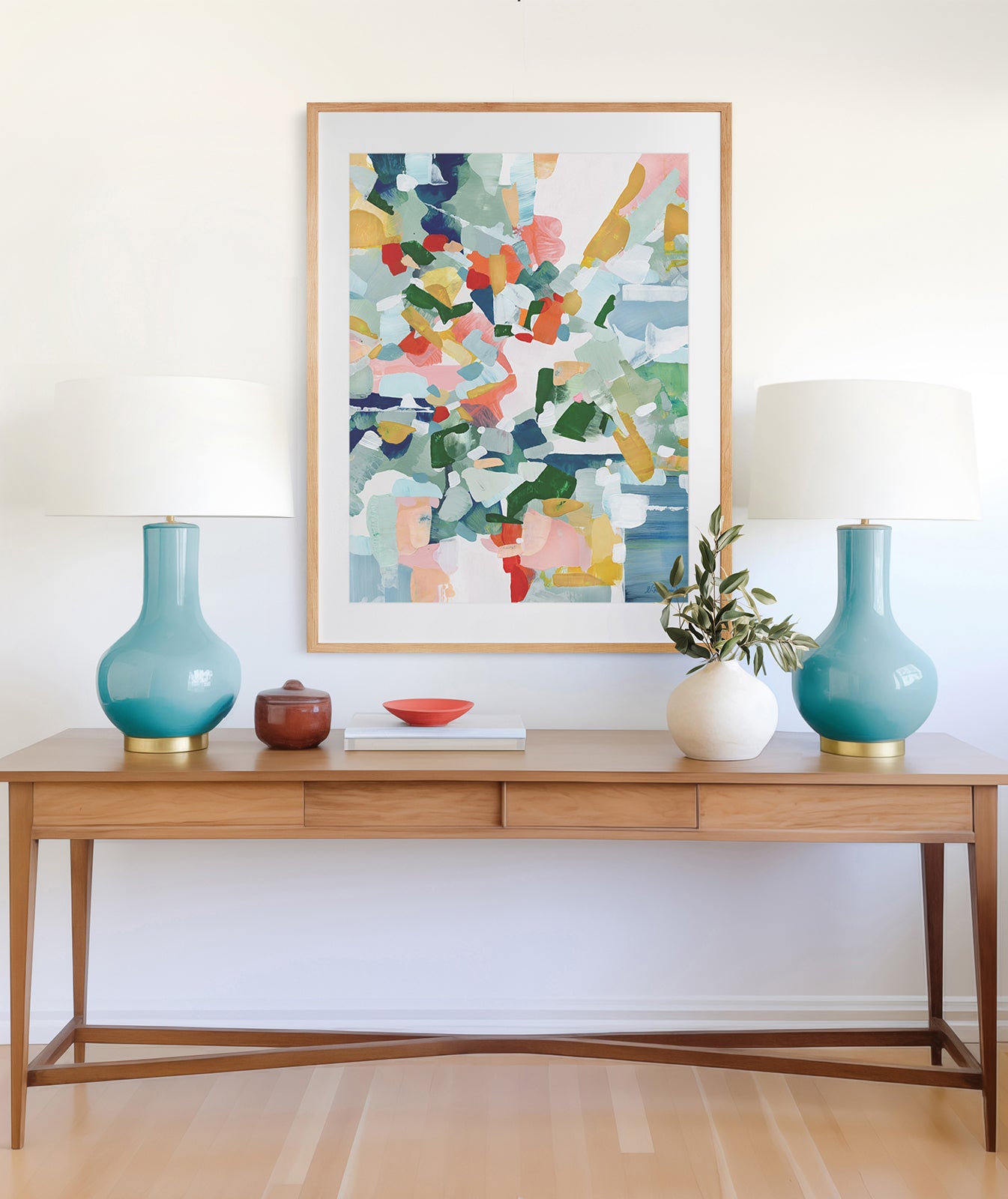 A large framed abstract art print in blue, red, yellow, pink and green displayed above a wooden table with two blue lamps.