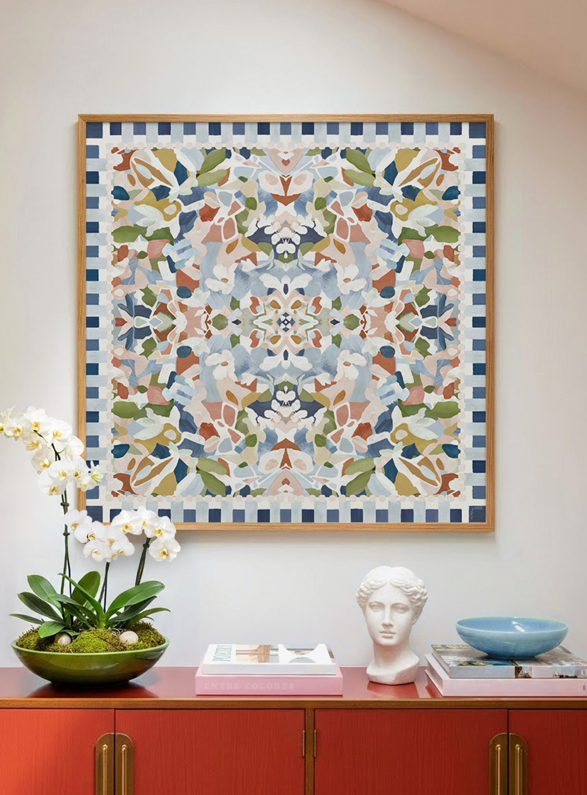 A framed square wall artwork with a colorful abstract floral and geometric pattern hangs on a light wall above a red cabinet displaying books, orchid flowers, a white head sculpture and a blue bowl. The artwork features a symmetrical multicolor design with blue, light pink, green, beige, orange, and white tones, surrounded by a blue-and-white check border.