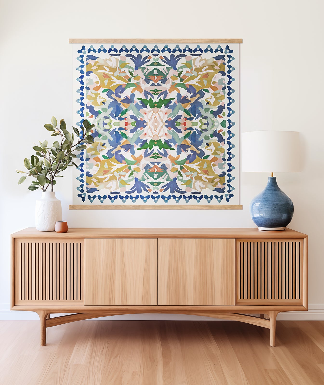 A colorful, abstract art print featuring shades of blue, yellow, green and pink hanging above a light wood cabinet with a blue lamp and white vase with branches, against a white wall.