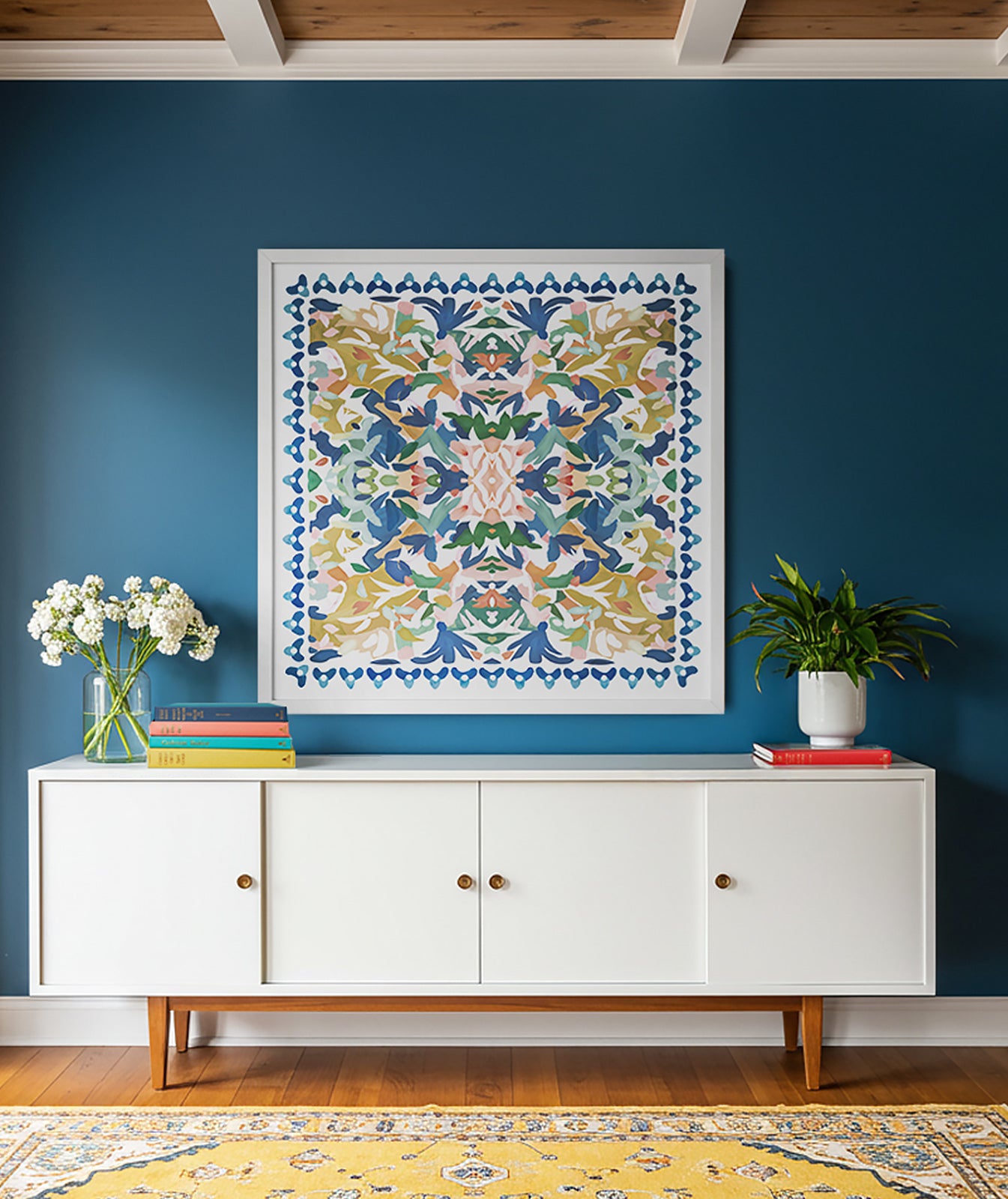  A mid-century modern space with blue walls, a white cabinet with books and flowers, above the cabinet is a large, square, abstract art print with vivid shades of blue, pinks and greens framed in white. There is a yellow, traditional rug on the floor.