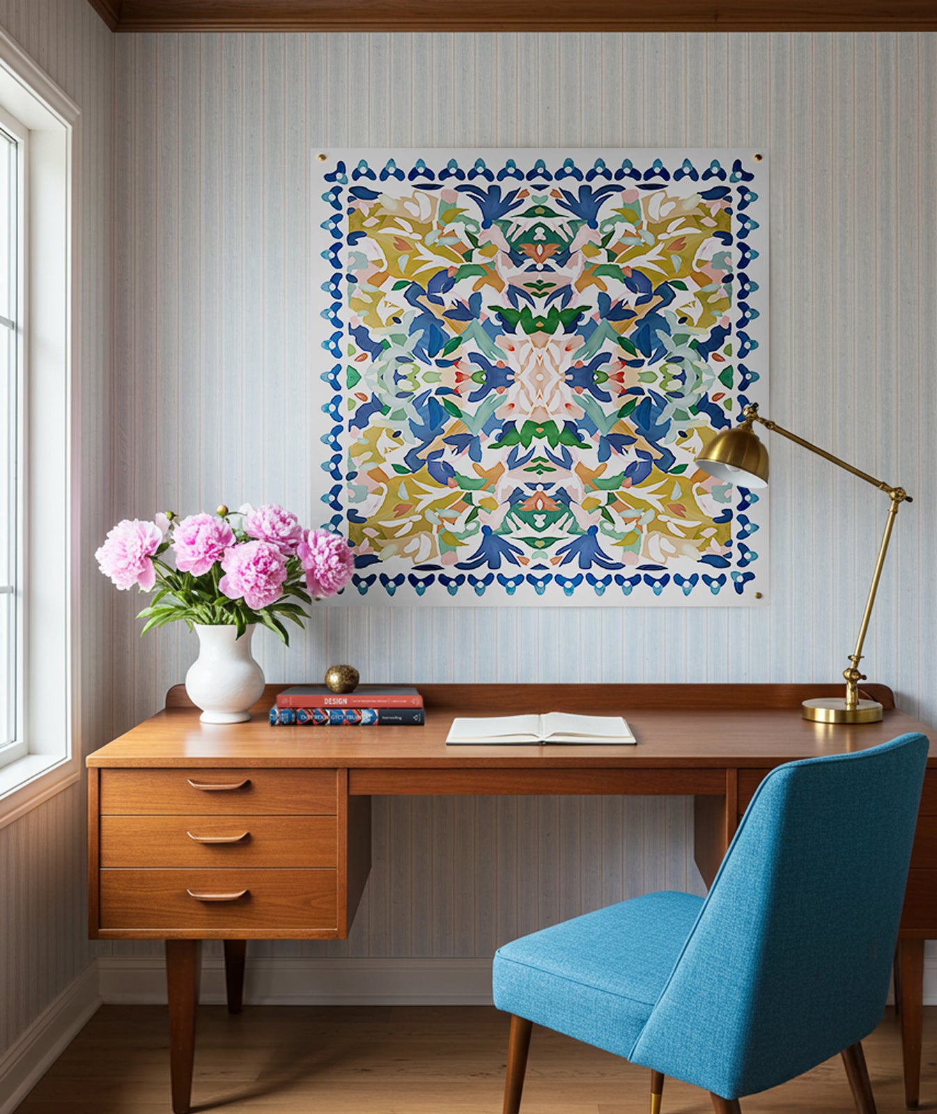 An abstract kaleidoscopic art print in blue, yellow, green and pink displayed on a wall above a wooden desk with a blue chair, a vase of pink flowers, and various desk items.
