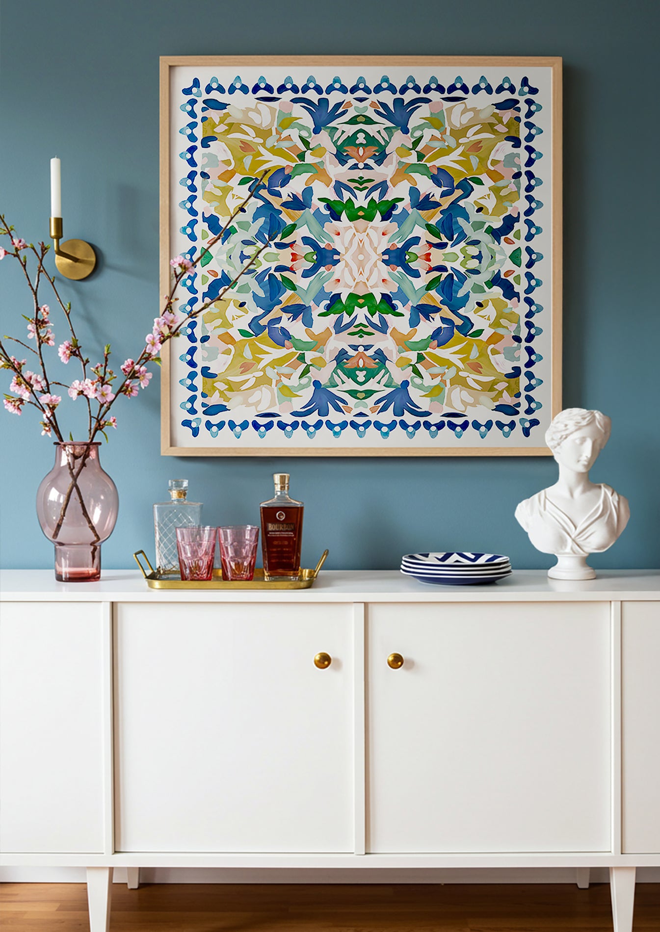 A framed, abstract kaleidoscopic art print in blue, yellow, green and pink displayed above a white cabinet against a blue wall.