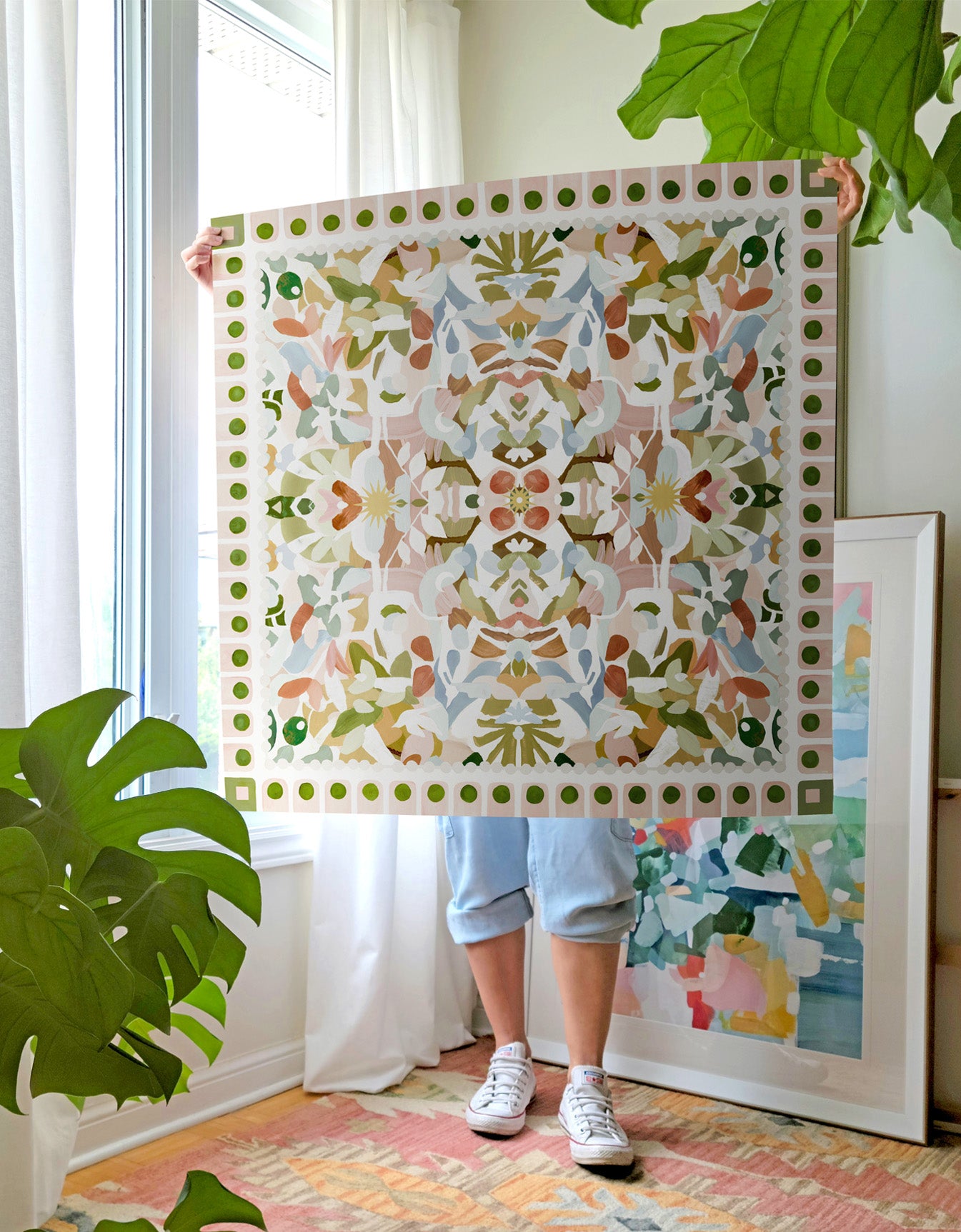 Person in a room with plants holding a large colorful symmetrical art print in light greens, pinks and blue tones.