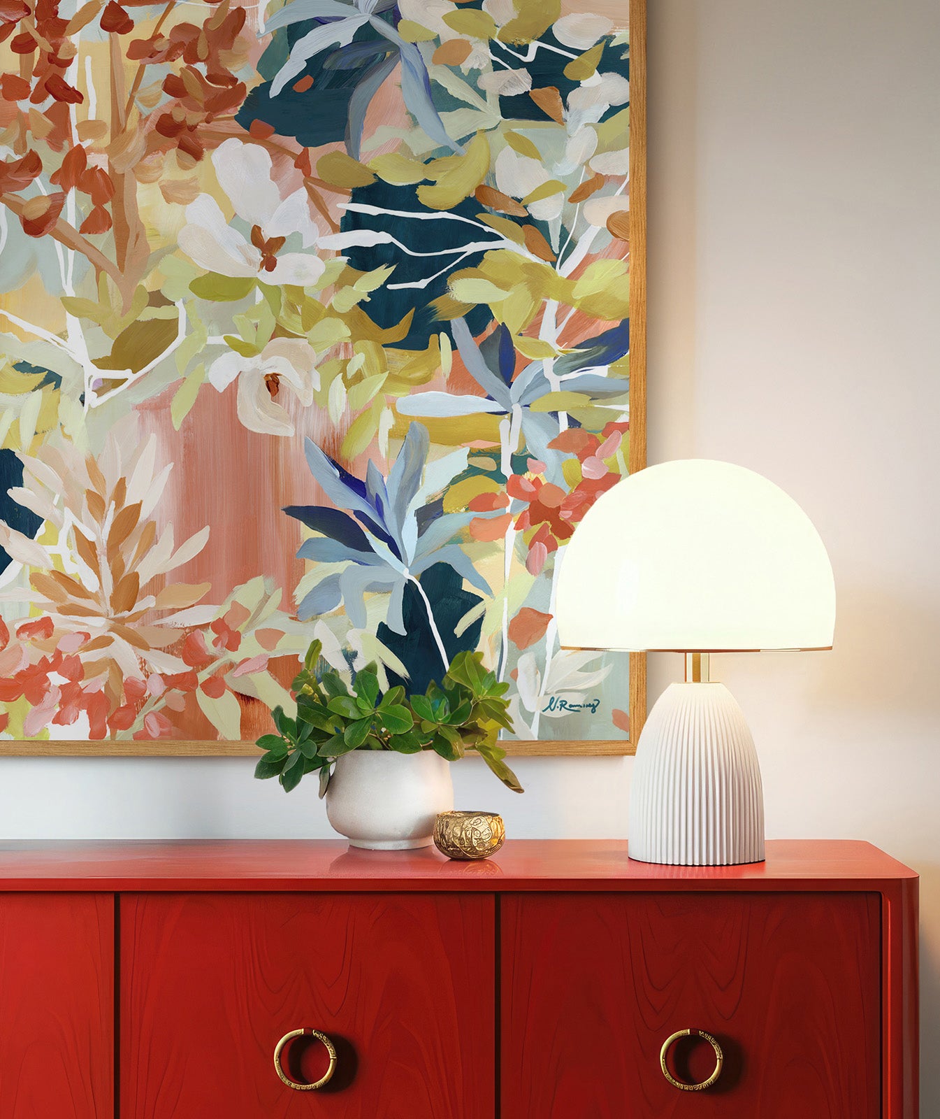 Closeup of a red cabinet with brass hardware, above it a large, floral, abstract art painting in red, blue and green colors, a white, mid century modern lamp next to it.