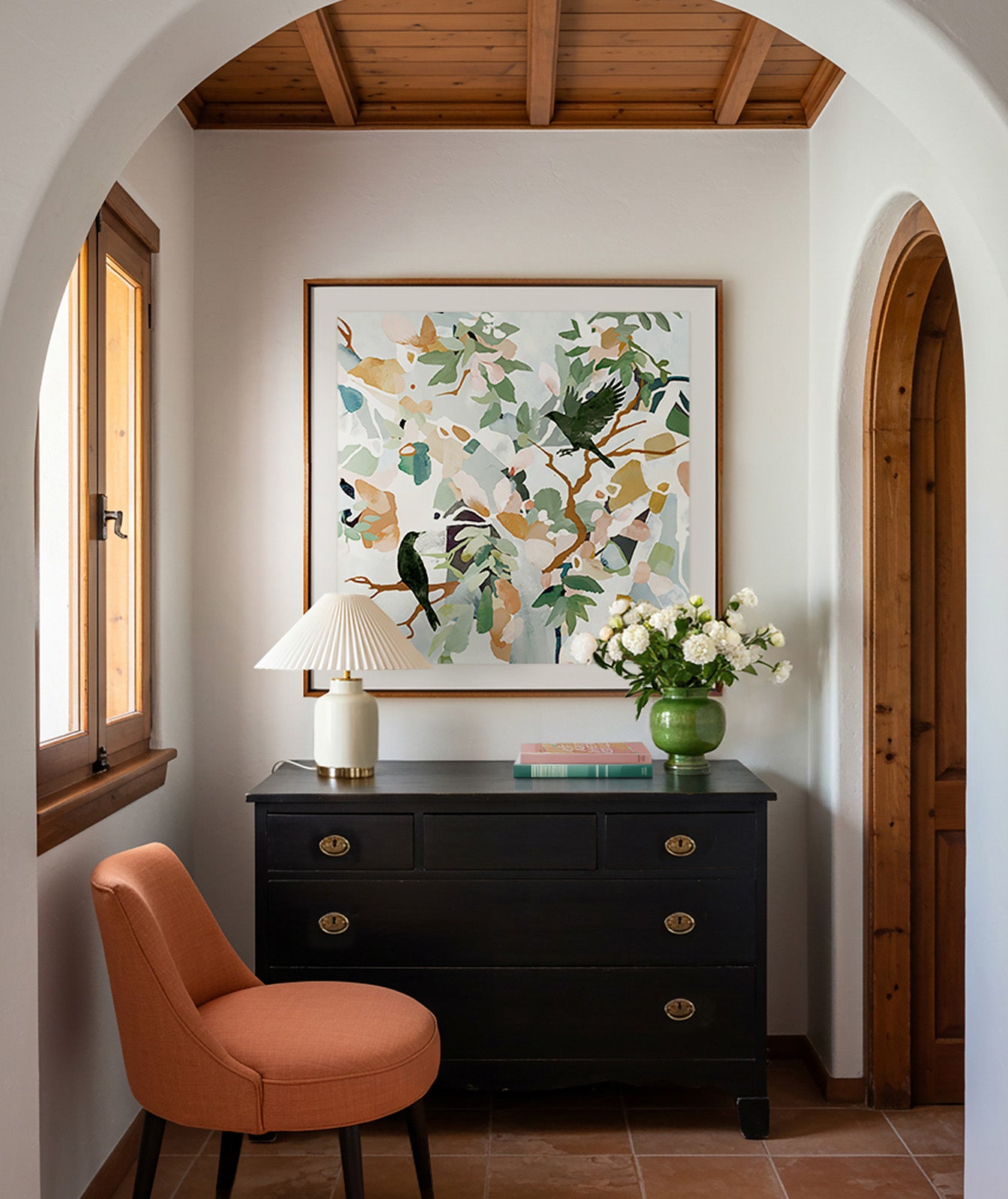 Narrow hallway with a black dresser, orange chair, and abstract colorful artwork featuring two black birds on a white wall.