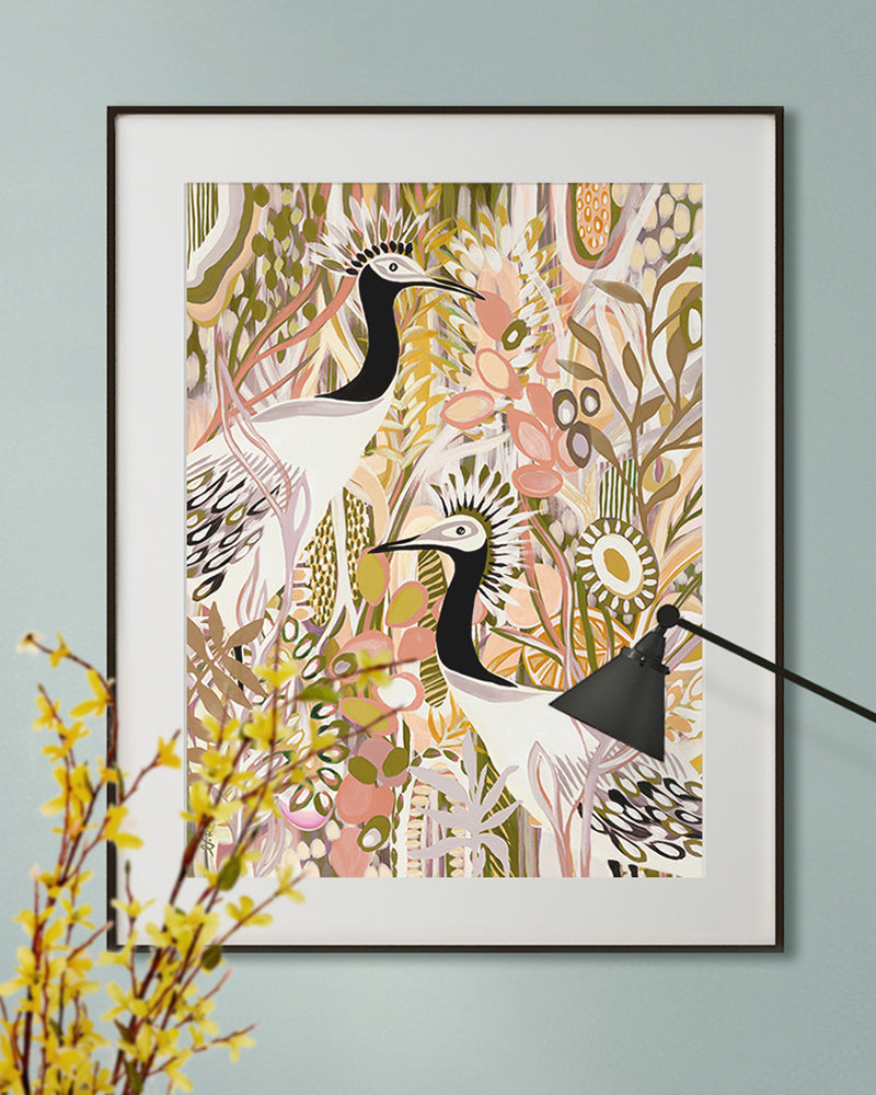 A framed fine art print displayed on a light blue wall featuring a floral design in pink, yellow and green along with two black and white birds.