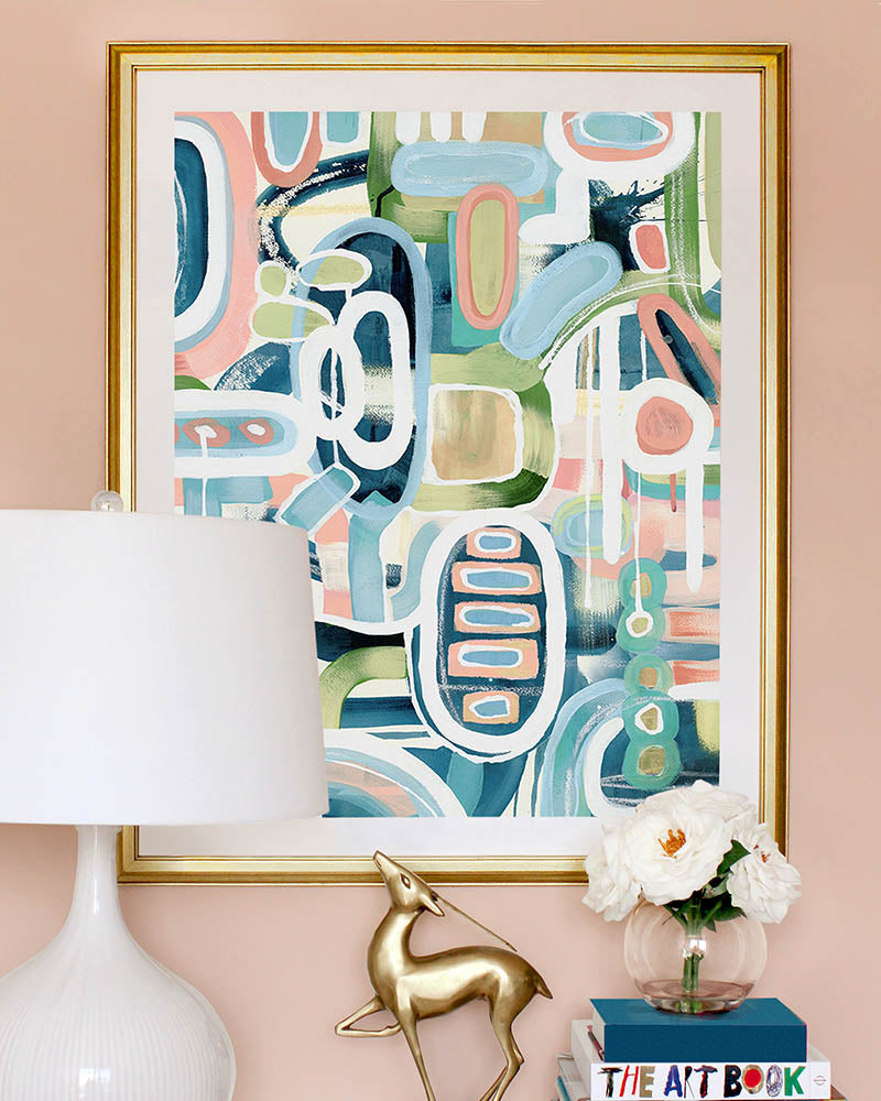 A framed, abstract art print with pink, green and blue colors, displayed alongside a white lamp, flowers and books.